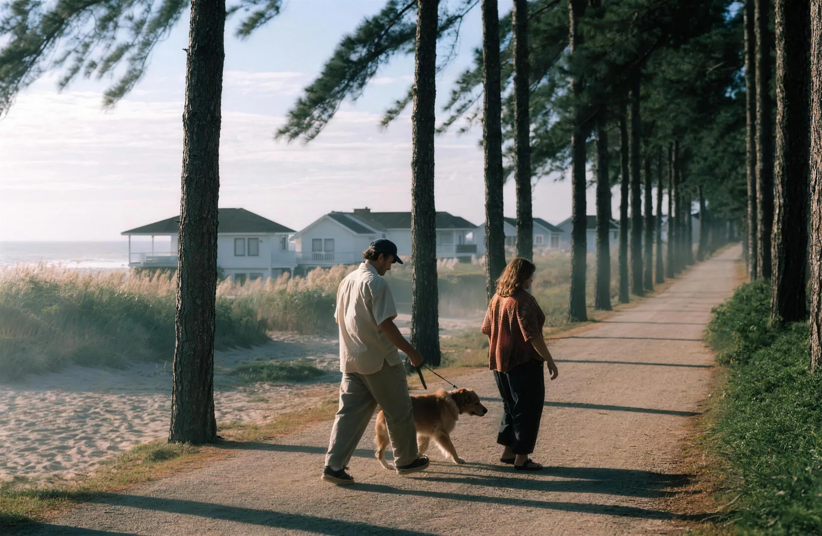 Couple walking a leashed golden retriever on a pine-shaded paved path with beach cottages behind